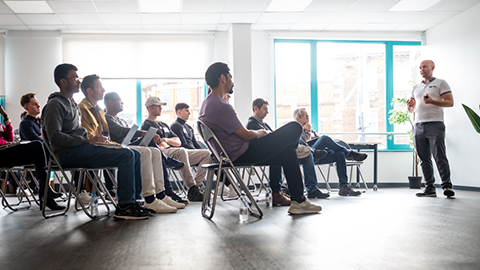 A room full of people sitting in chairs in casual clothing watching someone give a talk.